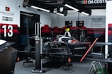 A Formula E race car is situated in a team garage. The car features prominent red and black coloring with the Porsche logo visible. Various equipment and tools for car maintenance and setup are scattered throughout the garage. On the wall, there are social media handles, driver information, and promotional content with the number 13 prominently displayed.