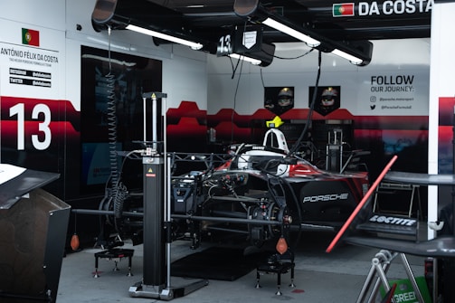 A Formula E race car is situated in a team garage. The car features prominent red and black coloring with the Porsche logo visible. Various equipment and tools for car maintenance and setup are scattered throughout the garage. On the wall, there are social media handles, driver information, and promotional content with the number 13 prominently displayed.