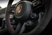 A close-up view of the interior of a car, featuring a steering wheel with the Porsche logo prominently displayed in the center. The steering wheel is detailed with black and red stitching. Two control knobs are visible, labeled 'Drive Mode' and 'ESC/TC'. The overall design conveys a modern, sporty aesthetic.