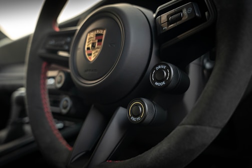 A close-up view of the interior of a car, featuring a steering wheel with the Porsche logo prominently displayed in the center. The steering wheel is detailed with black and red stitching. Two control knobs are visible, labeled 'Drive Mode' and 'ESC/TC'. The overall design conveys a modern, sporty aesthetic.