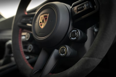 A close-up view of the interior of a car, featuring a steering wheel with the Porsche logo prominently displayed in the center. The steering wheel is detailed with black and red stitching. Two control knobs are visible, labeled 'Drive Mode' and 'ESC/TC'. The overall design conveys a modern, sporty aesthetic.