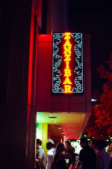 A neon sign displaying the word 'Zanzibar' in bright red and yellow, contrasting against a dark background. The sign is illuminated with floral or leaf-like patterns in blue. Below the sign, a group of people are standing under an entrance with soft ambient light, which creates a lively and engaging atmosphere.