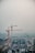 A construction site featuring two large cranes, one red and one yellow, towering over a developing building structure. The site appears busy with construction materials and equipment scattered throughout. The background is hazy, possibly due to fog or dust, adding a muted atmosphere to the scene.
