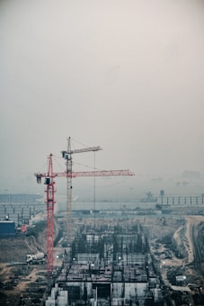 A construction site featuring two large cranes, one red and one yellow, towering over a developing building structure. The site appears busy with construction materials and equipment scattered throughout. The background is hazy, possibly due to fog or dust, adding a muted atmosphere to the scene.