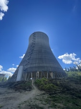 A large concrete cooling tower stands prominently against a clear blue sky. The tower has a hyperboloid structure with visible metal supports and openings near the base. Surrounding the tower is a grassy area with patches of brown and green vegetation. In the distance, there are trees and some power lines to the right.