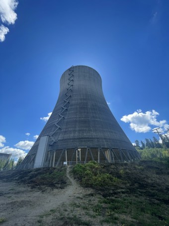 A large concrete cooling tower stands prominently against a clear blue sky. The tower has a hyperboloid structure with visible metal supports and openings near the base. Surrounding the tower is a grassy area with patches of brown and green vegetation. In the distance, there are trees and some power lines to the right.