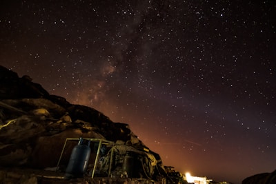 A star-filled night sky with the Milky Way visible in the background, creating a mesmerizing cosmic scene. The rocky foreground is dimly lit, with a prominent blue gas cylinder and some metal structures visible. A faint glow is seen on the horizon, suggesting distant artificial lights.