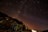 A star-filled night sky with the Milky Way visible in the background, creating a mesmerizing cosmic scene. The rocky foreground is dimly lit, with a prominent blue gas cylinder and some metal structures visible. A faint glow is seen on the horizon, suggesting distant artificial lights.