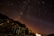 A star-filled night sky with the Milky Way visible in the background, creating a mesmerizing cosmic scene. The rocky foreground is dimly lit, with a prominent blue gas cylinder and some metal structures visible. A faint glow is seen on the horizon, suggesting distant artificial lights.