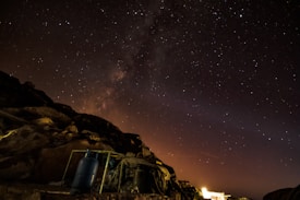 A star-filled night sky with the Milky Way visible in the background, creating a mesmerizing cosmic scene. The rocky foreground is dimly lit, with a prominent blue gas cylinder and some metal structures visible. A faint glow is seen on the horizon, suggesting distant artificial lights.