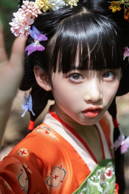 A young person dressed in traditional attire with an orange and green floral patterned garment. Their hair is intricately styled with brightly colored flowers and decorations. The lighting highlights their delicate facial features.
