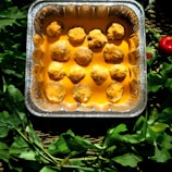 A tray of baked meatballs coated in a thick, creamy orange sauce is surrounded by fresh leafy greens and red vegetables.