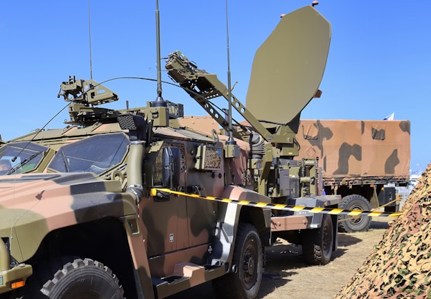 A military vehicle equipped with advanced communication or radar equipment is parked on a grassy area. The vehicle has a camouflage pattern and several antennas, including a large satellite dish mounted on a trailer. A camouflaged net is visible in the foreground, and another military vehicle is partially visible in the background.