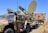 A military vehicle equipped with advanced communication or radar equipment is parked on a grassy area. The vehicle has a camouflage pattern and several antennas, including a large satellite dish mounted on a trailer. A camouflaged net is visible in the foreground, and another military vehicle is partially visible in the background.