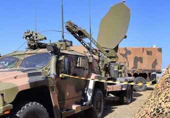 A military vehicle equipped with advanced communication or radar equipment is parked on a grassy area. The vehicle has a camouflage pattern and several antennas, including a large satellite dish mounted on a trailer. A camouflaged net is visible in the foreground, and another military vehicle is partially visible in the background.