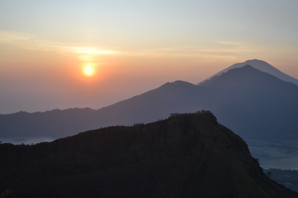 A serene landscape features a bright sunrise casting warm hues across the sky, with a gentle gradient from orange to blue. Two mountains, one prominent in the foreground and another smaller in the background, create a layered effect. Silhouettes of a few people are visible at the top of the near mountain, enhancing the sense of scale and exploration.
