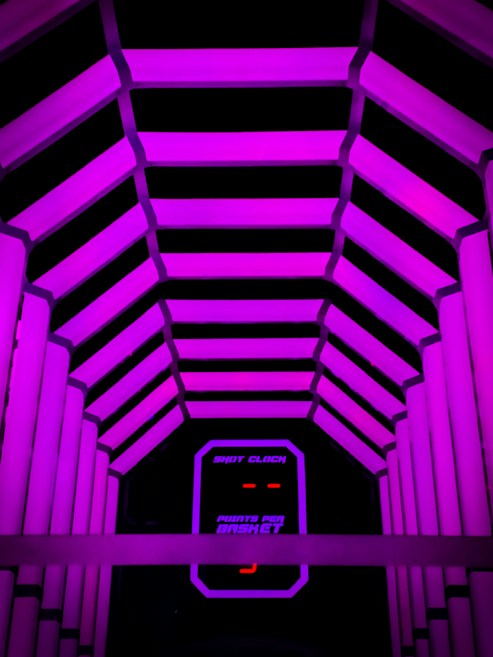 A tunnel-like structure illuminated with bright purple neon lights. The interior features parallel rows of glowing beams, creating a futuristic, geometric design. At the end of the tunnel, a digital display shows the text 'Shot Clock' and 'Points Per Basket' in a vibrant purple against a dark background.