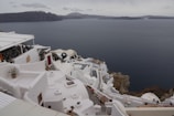 Whitewashed buildings with rounded roofs and terraces cascade down a hillside overlooking a calm sea, with rocky cliffs and distant islands visible in the misty background. Terracotta pots with small plants decorate some of the terraces.