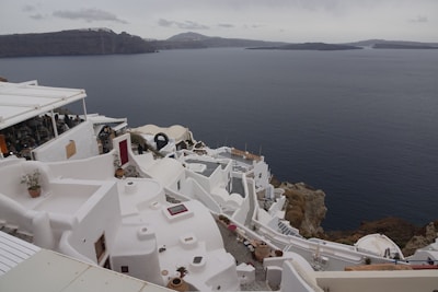 Whitewashed buildings with rounded roofs and terraces cascade down a hillside overlooking a calm sea, with rocky cliffs and distant islands visible in the misty background. Terracotta pots with small plants decorate some of the terraces.