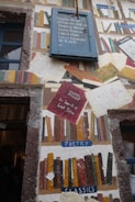 A wall mural features an eclectic assortment of painted books in various styles and colors, along with labeled sections such as 'Poetry' and 'Classics'. A blue sign with multilingual text offers books in different languages. The artwork mimics a library with a creative arrangement of book spines, titles, and texts on worn paper.