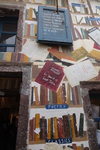 A wall mural features an eclectic assortment of painted books in various styles and colors, along with labeled sections such as 'Poetry' and 'Classics'. A blue sign with multilingual text offers books in different languages. The artwork mimics a library with a creative arrangement of book spines, titles, and texts on worn paper.