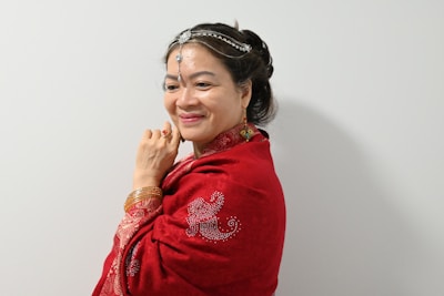 A woman in traditional attire stands against a plain background, wrapped in a rich red shawl adorned with intricate embroidery. She wears gold bangles, a decorative headpiece, and earrings, conveying a sense of cultural elegance.