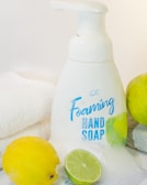 A bottle of foaming hand soap is surrounded by a lemon, a lime, and a soft white towel. The soap bottle is white with blue lettering and a label design featuring lemon imagery. The scene is clean and bright, with a citrus theme.