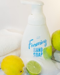 A bottle of foaming hand soap is surrounded by a lemon, a lime, and a soft white towel. The soap bottle is white with blue lettering and a label design featuring lemon imagery. The scene is clean and bright, with a citrus theme.