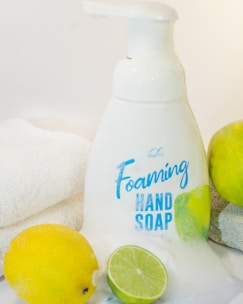 A bottle of foaming hand soap is surrounded by a lemon, a lime, and a soft white towel. The soap bottle is white with blue lettering and a label design featuring lemon imagery. The scene is clean and bright, with a citrus theme.