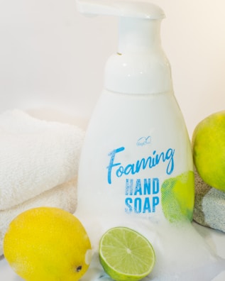 A bottle of foaming hand soap is surrounded by a lemon, a lime, and a soft white towel. The soap bottle is white with blue lettering and a label design featuring lemon imagery. The scene is clean and bright, with a citrus theme.