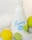A bottle of foaming hand soap is surrounded by a lemon, a lime, and a soft white towel. The soap bottle is white with blue lettering and a label design featuring lemon imagery. The scene is clean and bright, with a citrus theme.