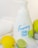 A bottle of foaming hand soap is surrounded by a lemon, a lime, and a soft white towel. The soap bottle is white with blue lettering and a label design featuring lemon imagery. The scene is clean and bright, with a citrus theme.