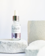 A dropper bottle labeled 'nuface Nu Glow' is positioned on a textured white stone surface with additional stone blocks in the background. The bottle contains a skin serum with a clear glass body and a metallic cap.