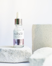 A dropper bottle labeled 'nuface Nu Glow' is positioned on a textured white stone surface with additional stone blocks in the background. The bottle contains a skin serum with a clear glass body and a metallic cap.