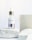 A dropper bottle labeled 'nuface Nu Glow' is positioned on a textured white stone surface with additional stone blocks in the background. The bottle contains a skin serum with a clear glass body and a metallic cap.