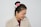 A woman with her hair styled in a bun adorned with pink and white flowers. She is wearing a light beige coat with a collar and gold earrings. The background is a plain light blue wall. She is looking downwards with a smile on her face.