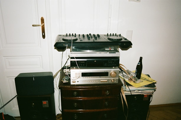 A DJ setup with various audio equipment stacked on a wooden cabinet, including a mixer, turntable, and amplifier. There is a beer bottle and a stack of vinyl records on the side. The equipment is surrounded by speakers and cables.