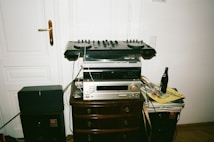 A DJ setup with various audio equipment stacked on a wooden cabinet, including a mixer, turntable, and amplifier. There is a beer bottle and a stack of vinyl records on the side. The equipment is surrounded by speakers and cables.