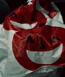 A crinkled surface featuring a red and white jack-o'-lantern pattern with prominent triangular eyes and a wide, curved smile. The material appears to be plastic or a similar synthetic fabric, giving it a slightly shiny texture.