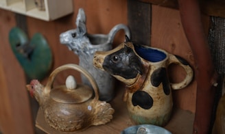 A rustic shelf displays handcrafted pottery including a jug with a cow head handle, a pitcher with a llama face, and a teapot shaped like a chicken.