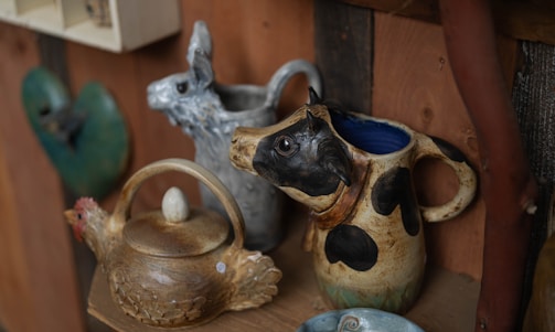 A rustic shelf displays handcrafted pottery including a jug with a cow head handle, a pitcher with a llama face, and a teapot shaped like a chicken.