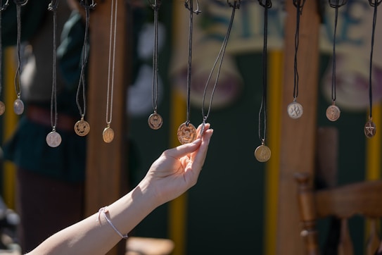 A hand reaches up to touch a hanging necklace with a circular pendant among a series of similar necklaces. Each necklace is suspended from black cords. The background features blurred shapes and colors, suggesting an outdoor setting possibly at a market or fair.