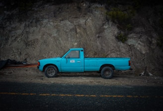 A vintage turquoise pickup truck is parked against a rocky and earthy terrain. The truck is old and a bit worn, with a flatbed and noticeable rust on its wheels. The background consists of rugged earth and stones, suggesting an off-road or remote location.