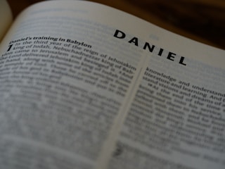 A close-up of an open book, specifically the page showing the beginning of the Book of Daniel. The heading 'DANIEL' is prominently displayed in bold font, with the text underneath detailing the onset of Daniel's training in Babylon.