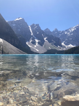 A serene alpine lake with crystal clear water reflecting a backdrop of majestic snow-capped mountains. The rocky shoreline is visible beneath the water's surface, and the sky above is clear and bright.