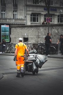 A worker in bright orange reflective clothing pushes a cart loaded with garbage bags down a city street. The background features bicycles lined up against a railing, pedestrians walking by, and large buildings with stone facades. A digital billboard and a no parking sign are visible, adding to the urban setting.