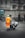 A worker in bright orange reflective clothing pushes a cart loaded with garbage bags down a city street. The background features bicycles lined up against a railing, pedestrians walking by, and large buildings with stone facades. A digital billboard and a no parking sign are visible, adding to the urban setting.