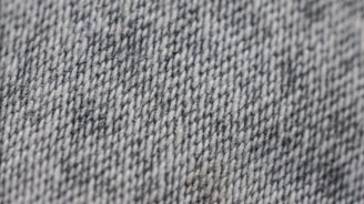 A close-up view of a textured fabric surface with a woven pattern, primarily consisting of gray and white threads. The texture appears coarse, suggesting a sturdy material.