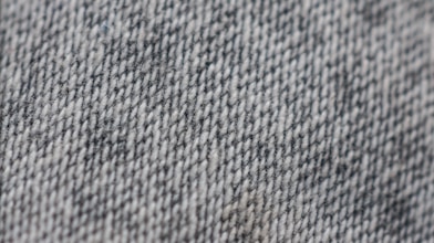 A close-up view of a textured fabric surface with a woven pattern, primarily consisting of gray and white threads. The texture appears coarse, suggesting a sturdy material.