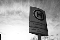 A black and white photograph depicting a public transportation sign with the letter 'H' prominently displayed. Below the letter are location names and the bus route number 147 to S-Bhf Ostbahnhof. The sign is set against a cloudy sky with bare tree branches visible at the bottom.